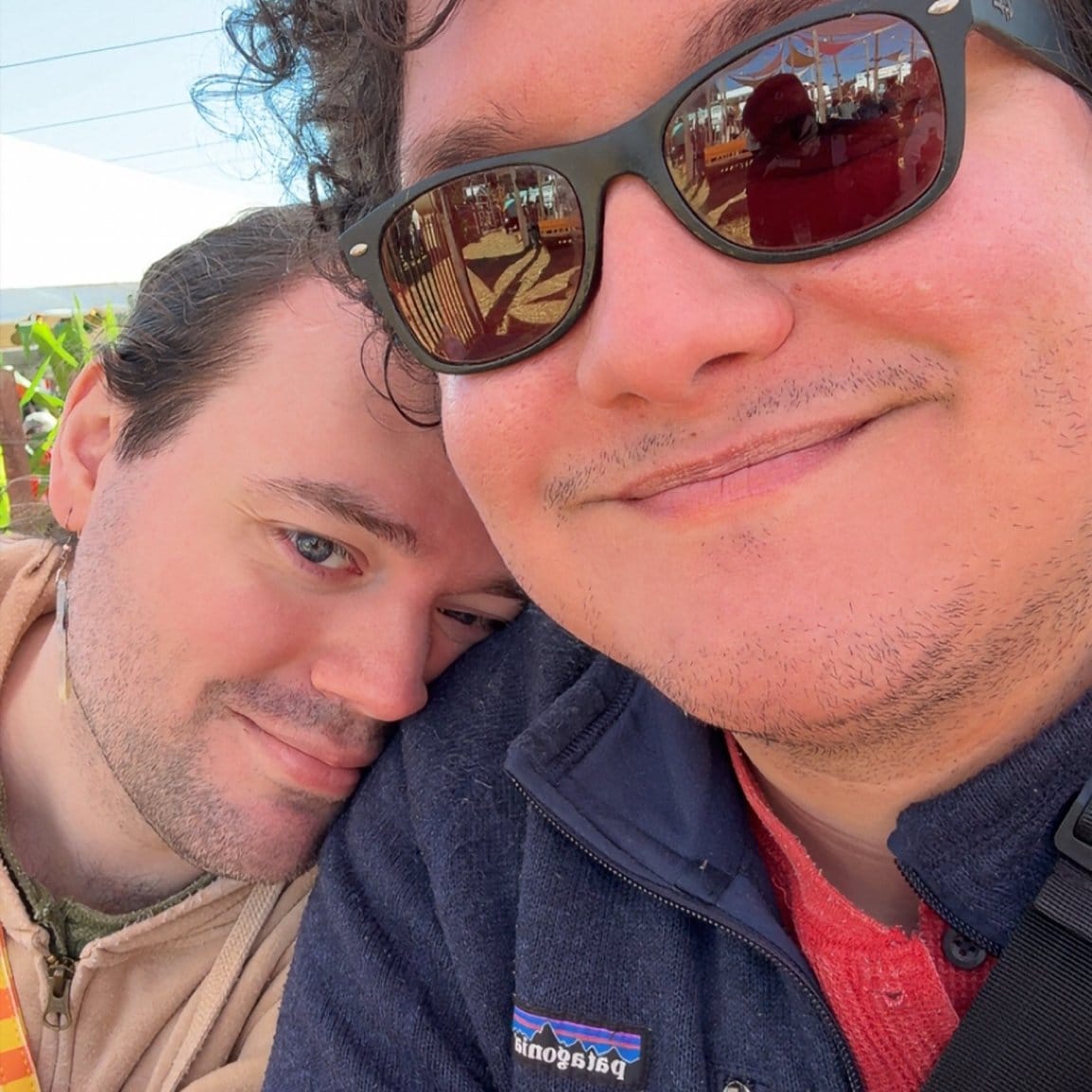 A picture of me and my husband. I’m wearing an orange Henley under a dark blue Patagonia zip up sweater and wearing glasses. I have curly black hair and I’m smiling at the camera. My husband is on my shoulder looking at the camera. They have a green Henley under a tan hoodie. They have long hair and are smiling.